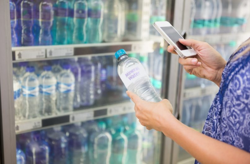 Woman comparing the price of a bottle of water with her phone at the supermarket